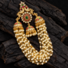 Gold-tone peacock jhumka hair ornament with green centre stone, ruby-red accents, three decorative jhumkas and layered white pearl chains.